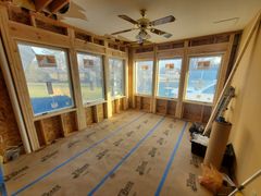 Unfinished sunroom with exposed framing, large windows, ceiling fan, and protective floor covering