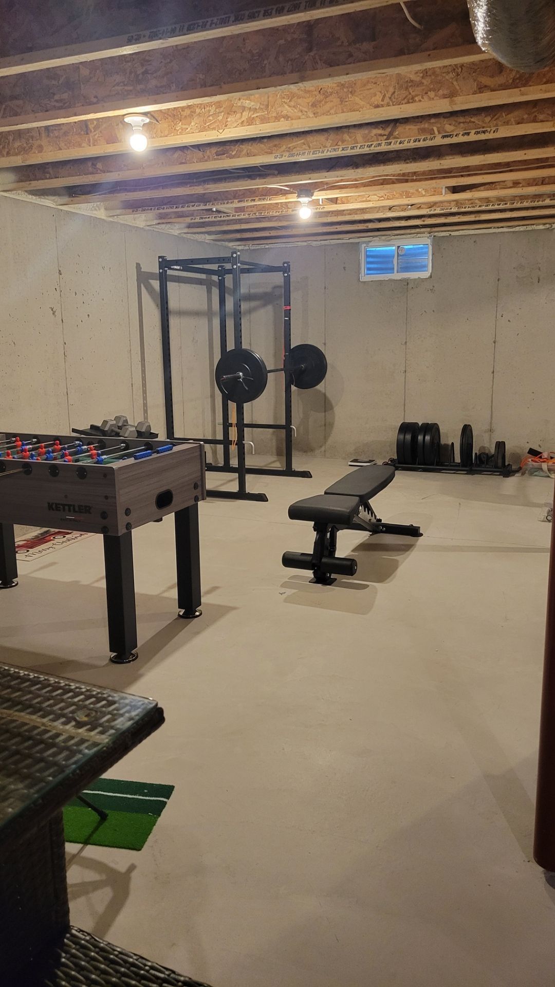 Basement home gym with weight bench, cable machine, foosball table, and exposed ceiling beams