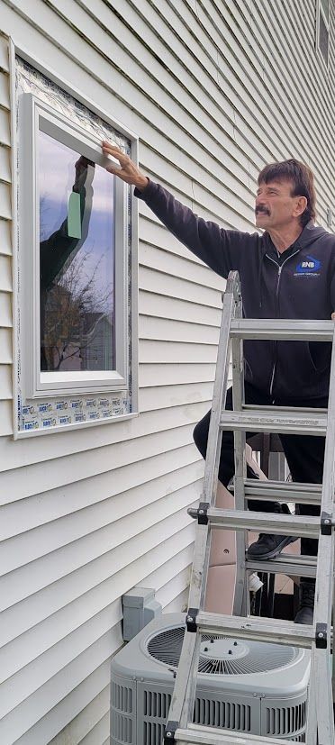Man on a ladder painting exterior siding beside a window and air conditioner unit