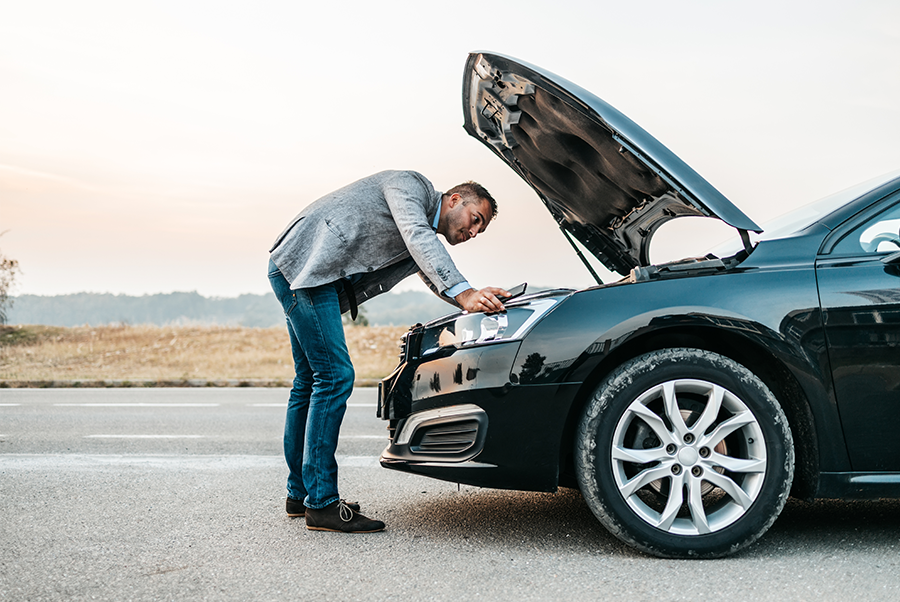 Man leaning over open hood of black car on roadside, inspecting engine near desert landscape