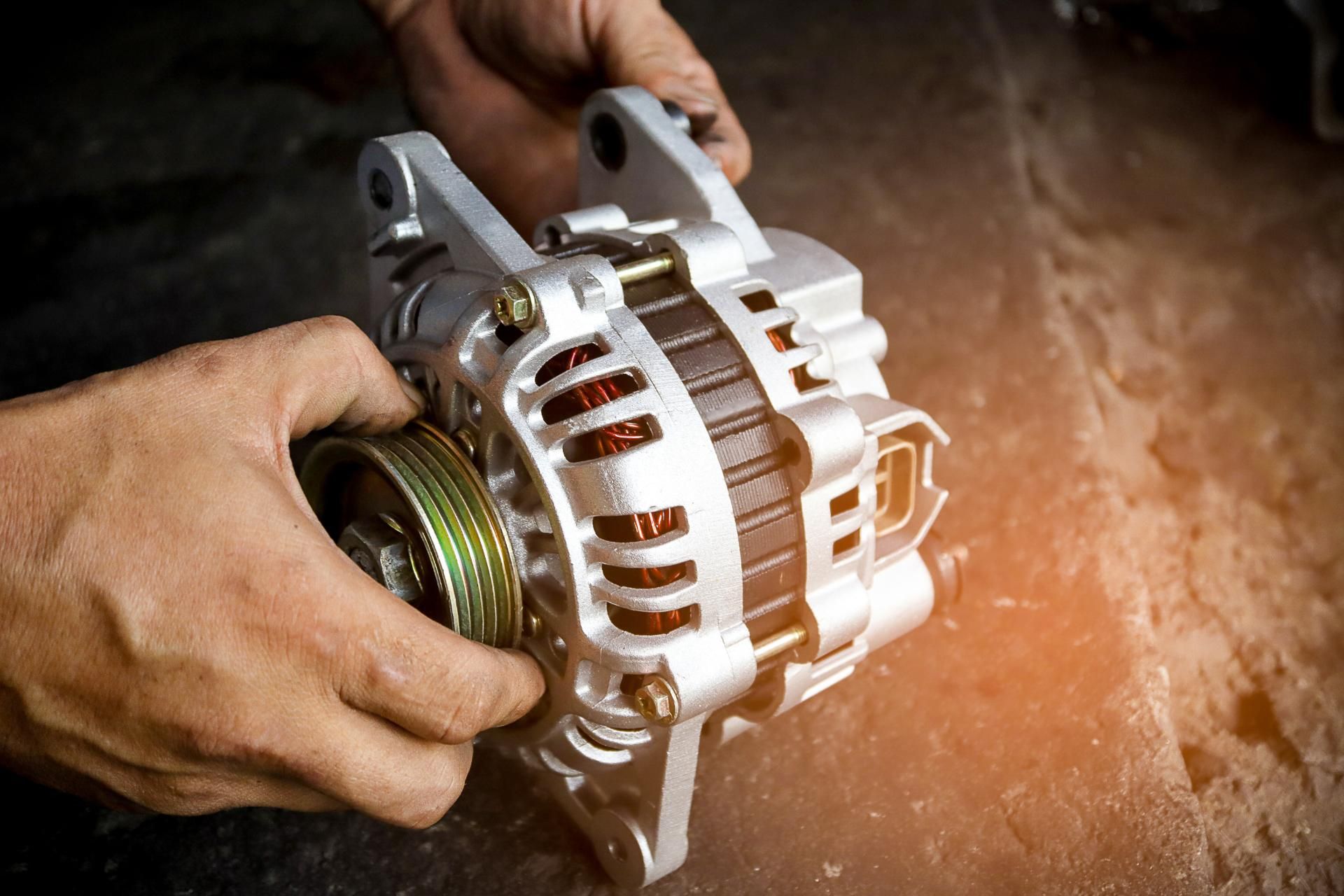 Hands holding a silver car alternator during repair on a workbench