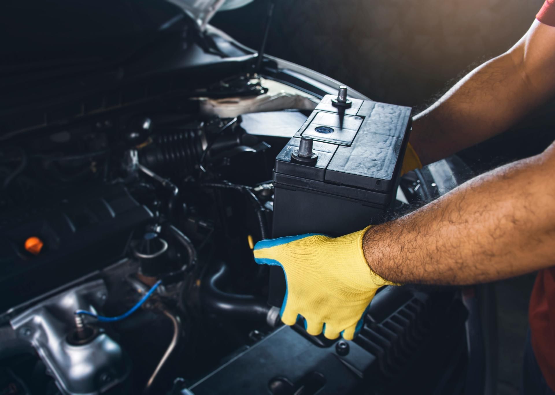 Person in yellow gloves replacing a car battery under the hood of an open vehicle