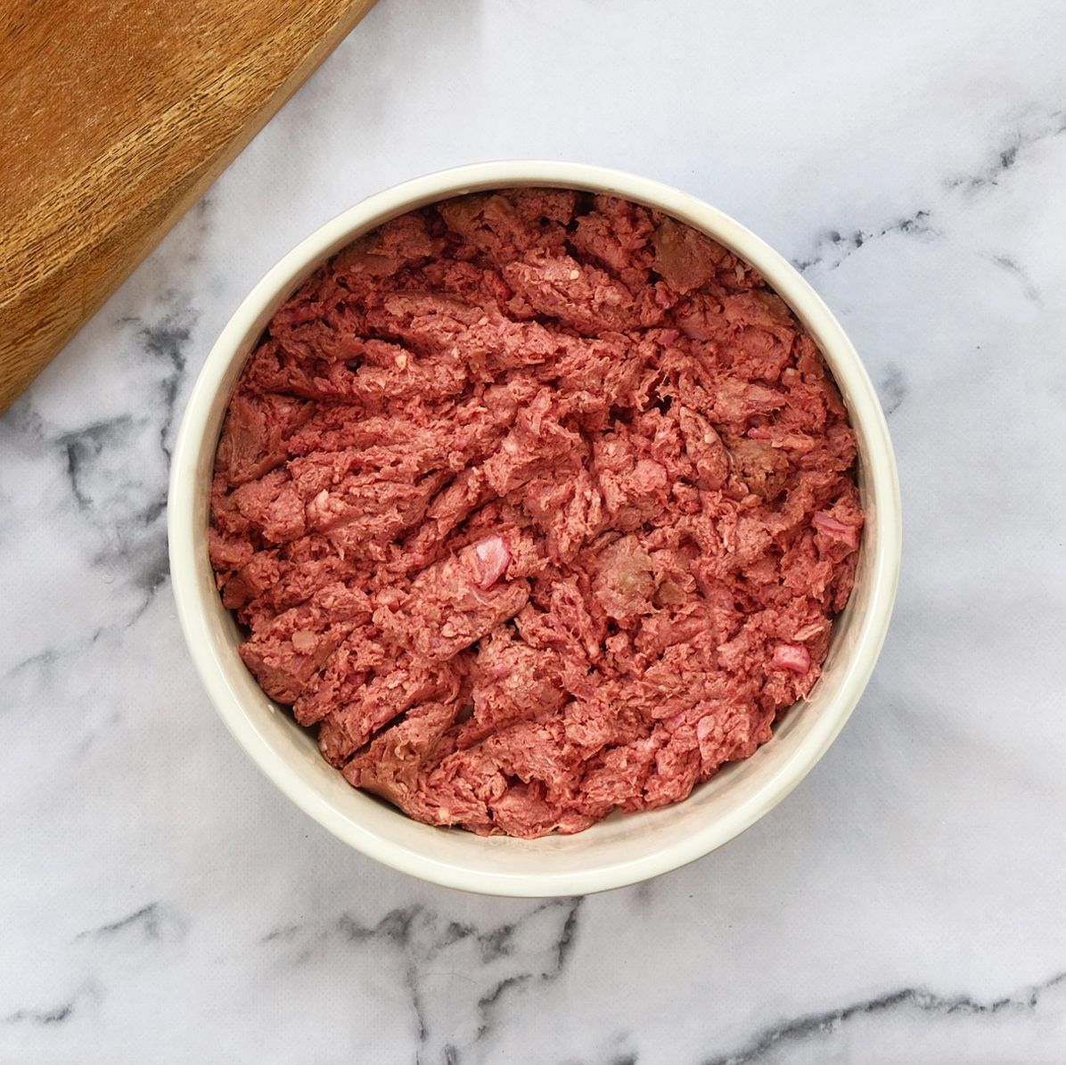 Bowl of raw, ground meat on a white and gray marbled surface, with a wooden board in the corner.
