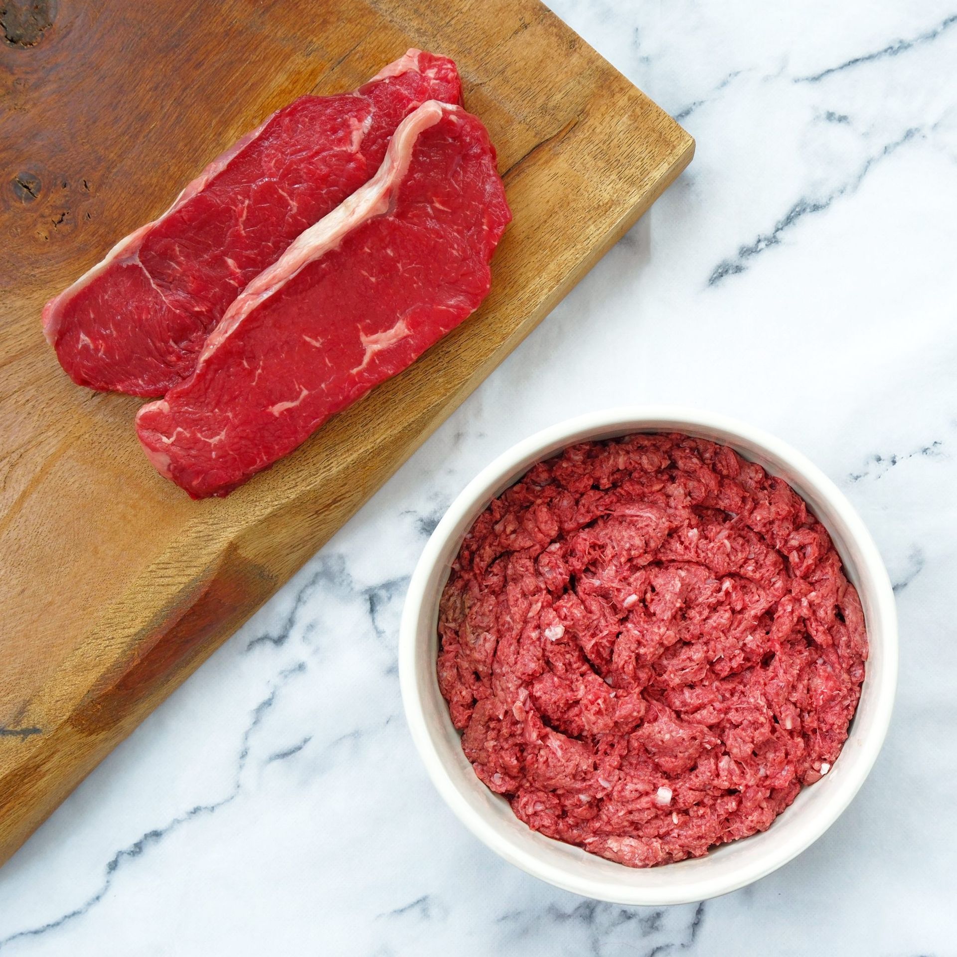 Two raw steaks on a wooden board next to a bowl of ground meat.