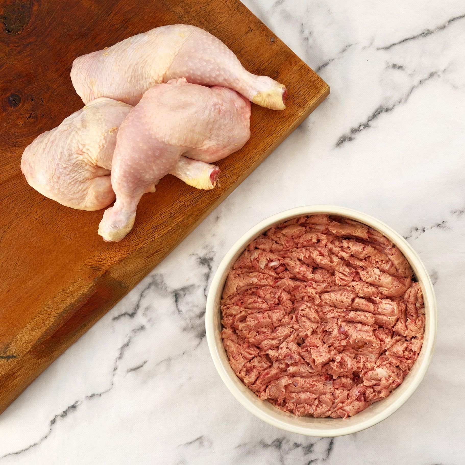 Raw chicken legs on a wooden board next to a bowl of ground raw meat on a marble surface.