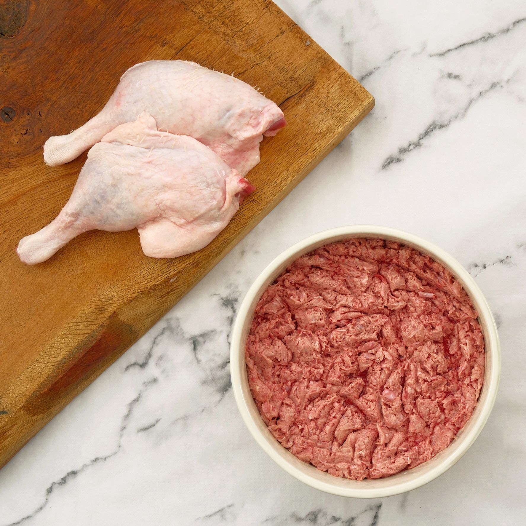 Duck legs on a wooden cutting board next to a bowl of raw, ground duck meat.