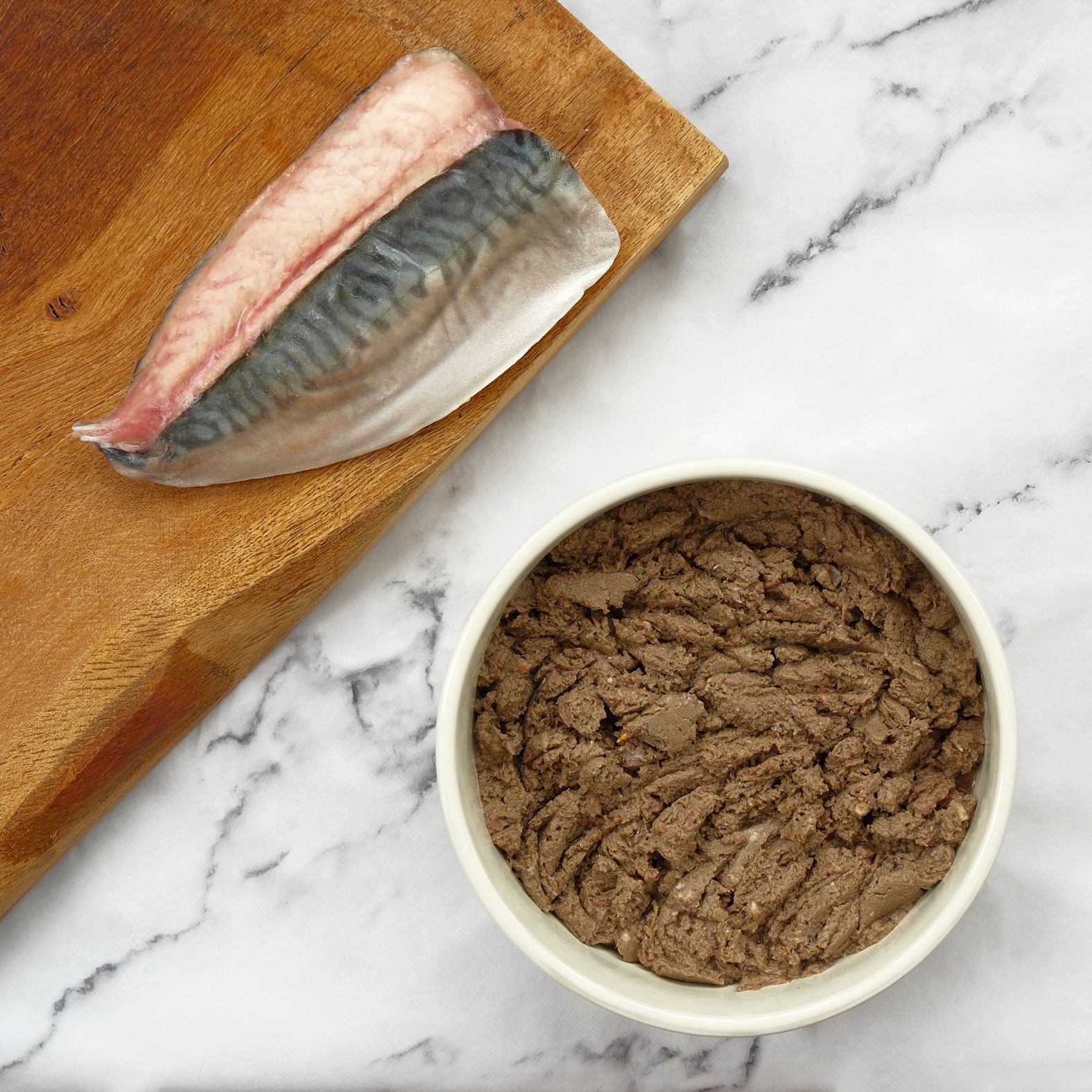 Mackerel fillet on a wooden board, with a bowl of brown, pâté-like substance on a marble surface.