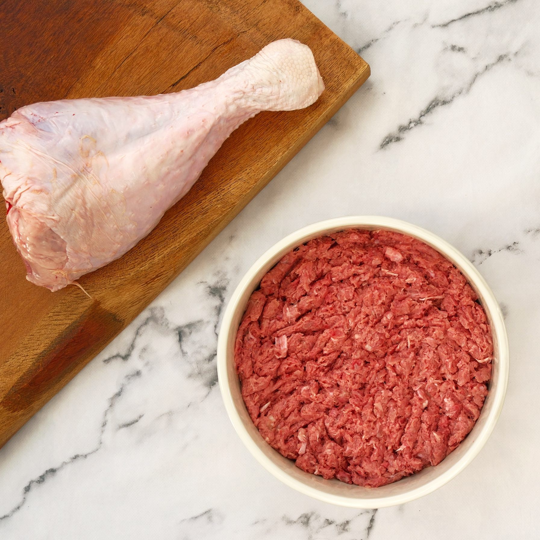 Turkey leg on a wood cutting board, next to a bowl of raw ground meat, on a white marble surface.