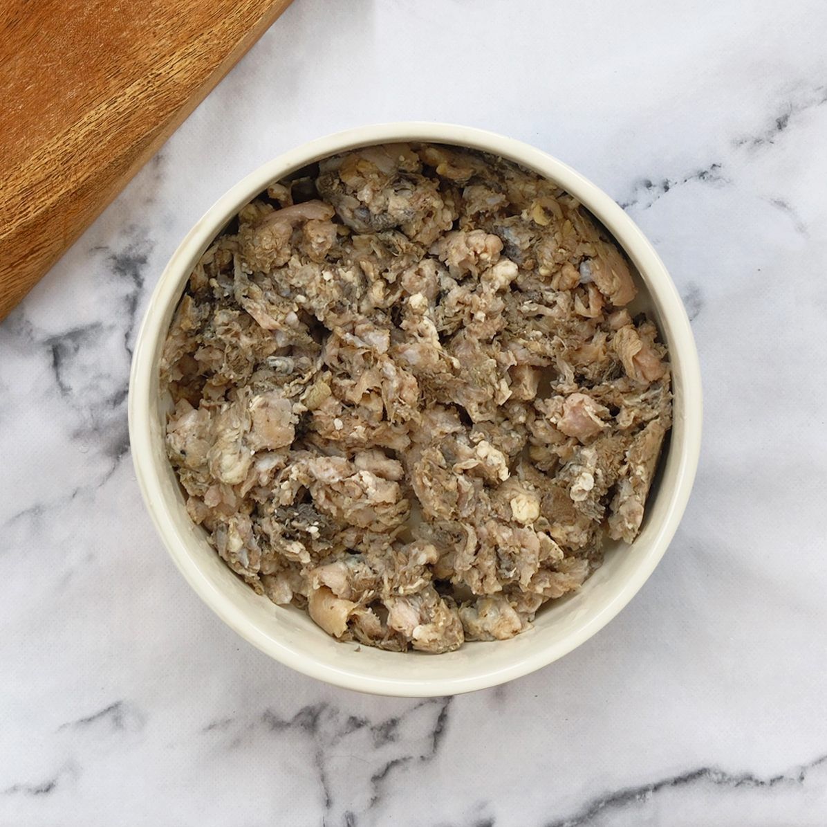 A white bowl filled with chunky, brown, minced wet pet food sits on a white marble surface next to a wooden object.