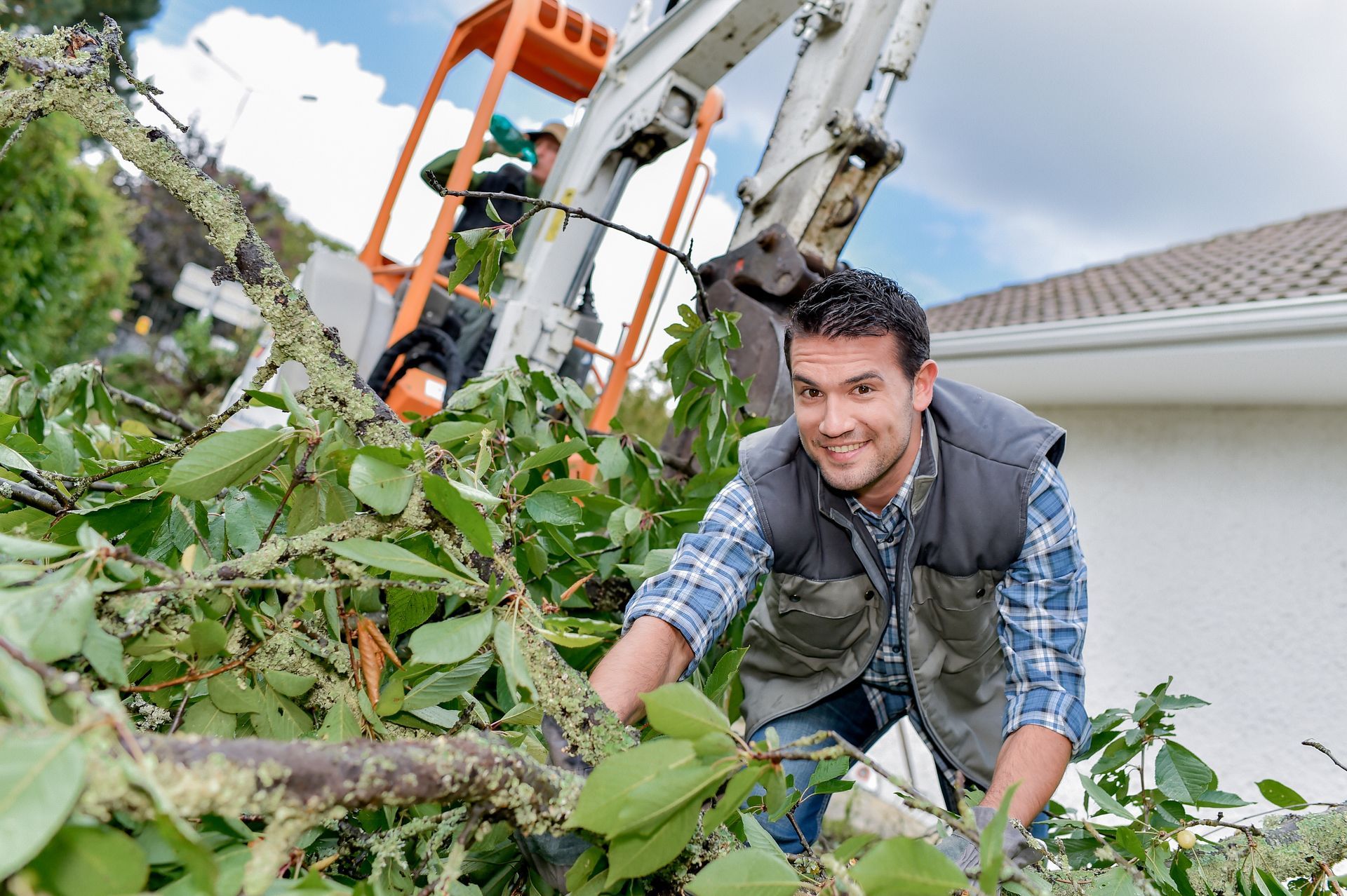 A person uses a chainsaw to cut down a tree with branches and leaves scattered on the ground. Person clearing tree branches and yard debris near an excavator at a residential property.