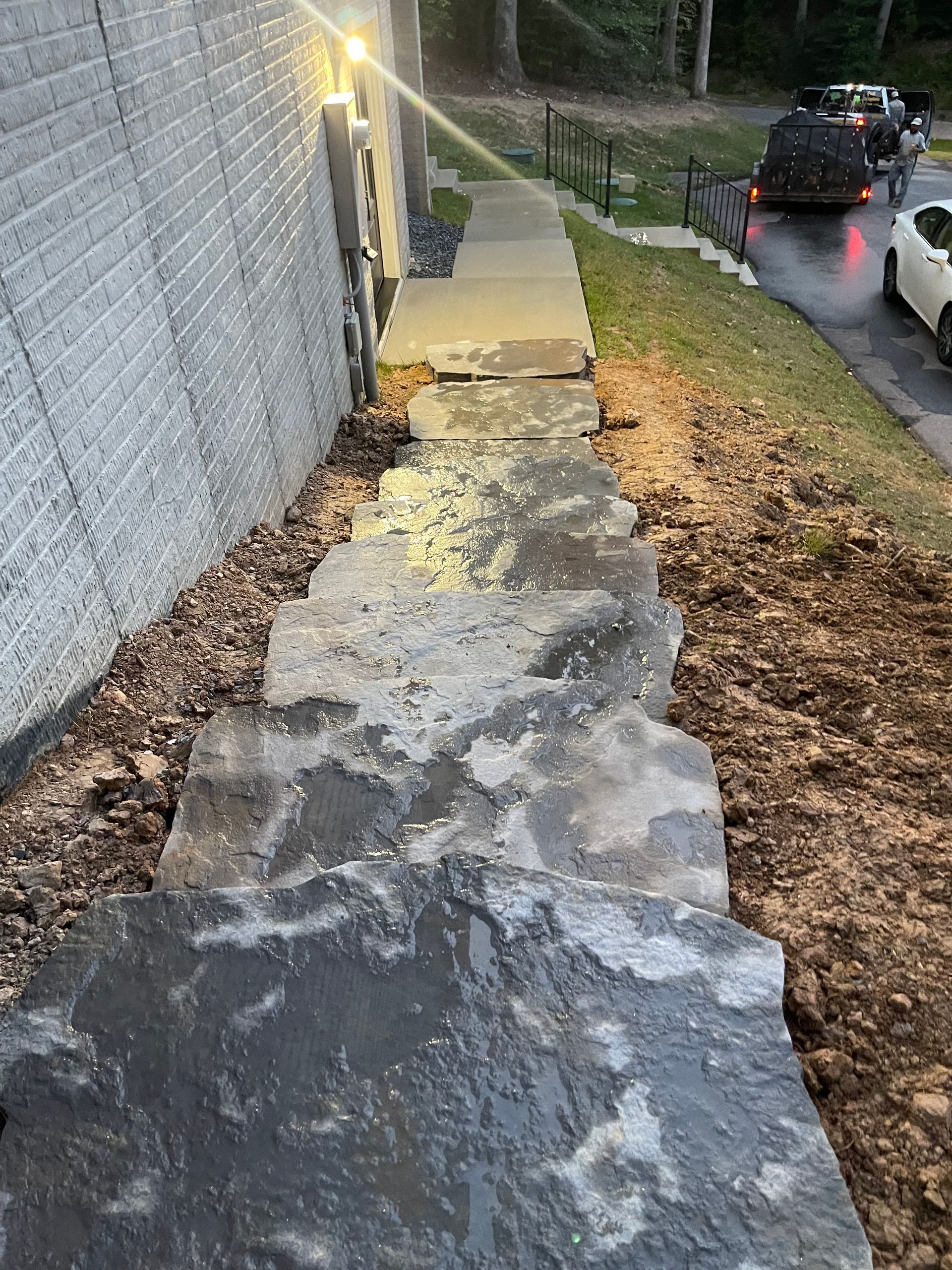 A set of uneven, natural stone steps along a light-colored brick wall, leading down to a street with cars nearby.