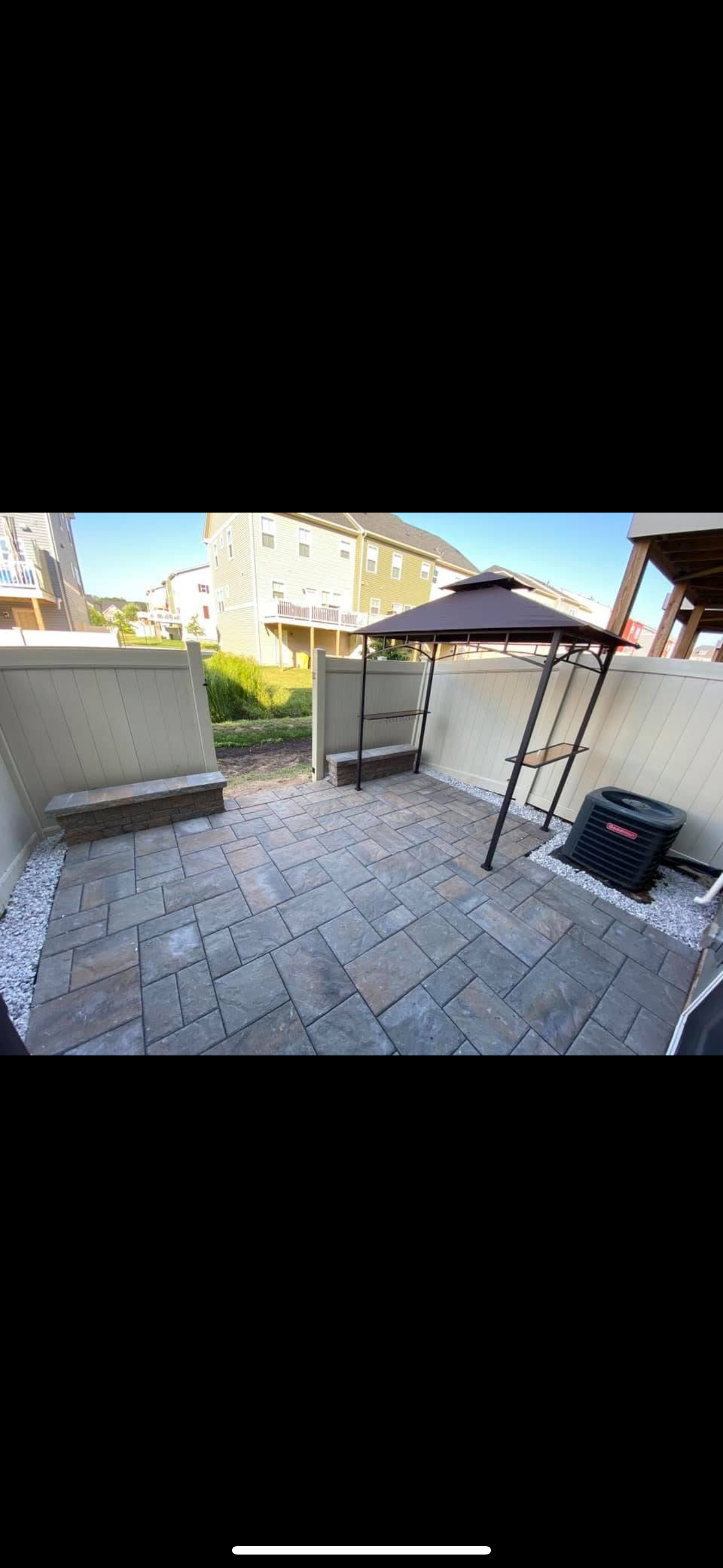 A stone-paved patio with a small gazebo, white privacy walls, a bench, and an outdoor HVAC unit on a sunny day.