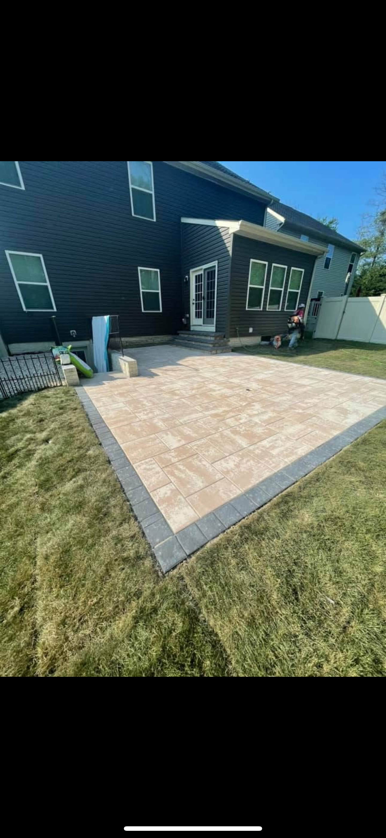 A newly installed rectangular patio with beige pavers and a dark gray border outside a dark blue residential house.