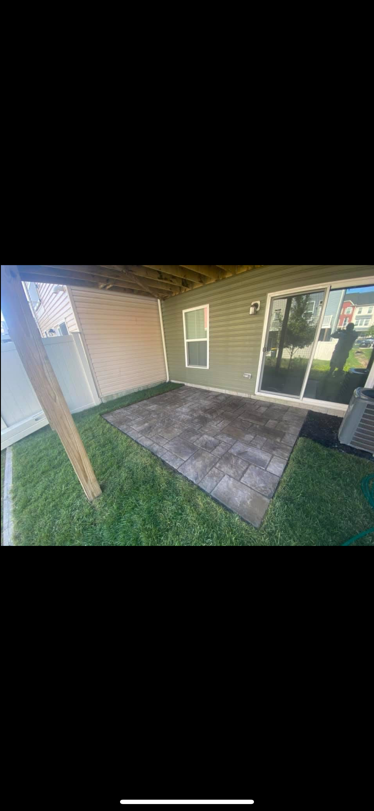A patio with dark pavers sits in a small backyard under an elevated wooden deck, adjacent to a house with sliding doors.