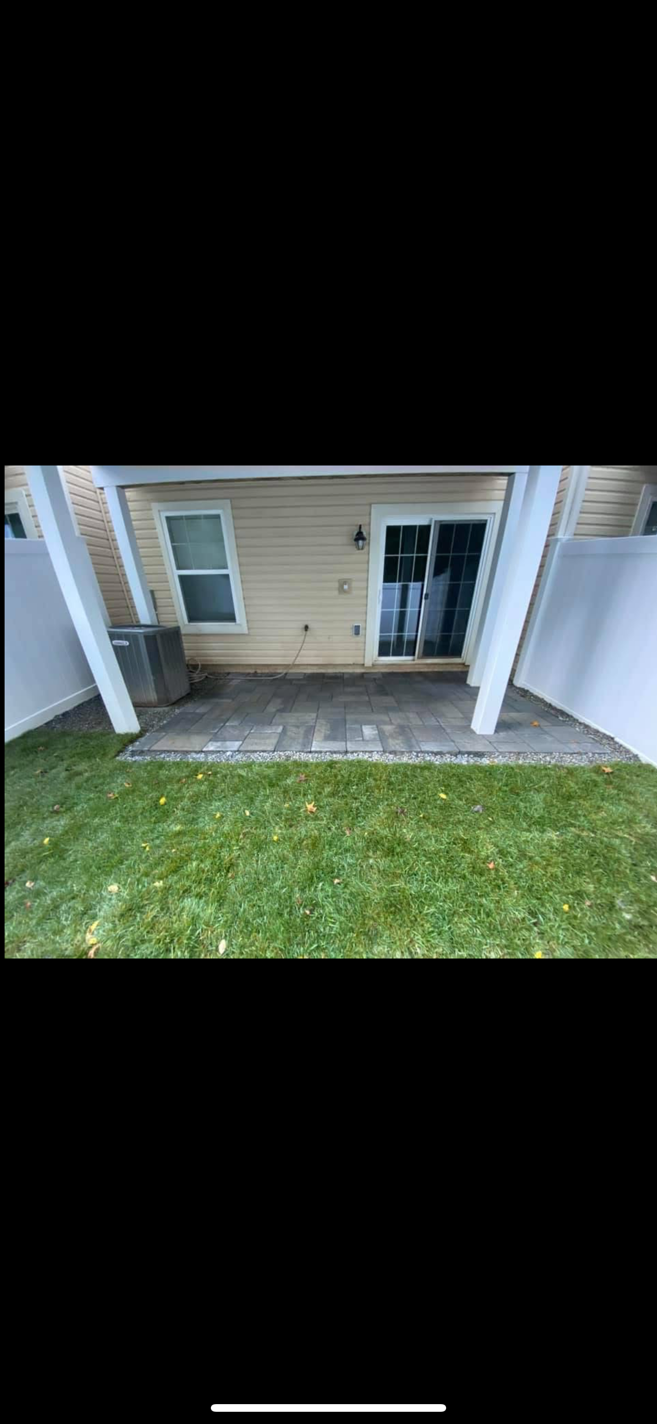A tan-sided house exterior features a small stone patio under a roof overhang, leading to a sliding glass door and yard.
