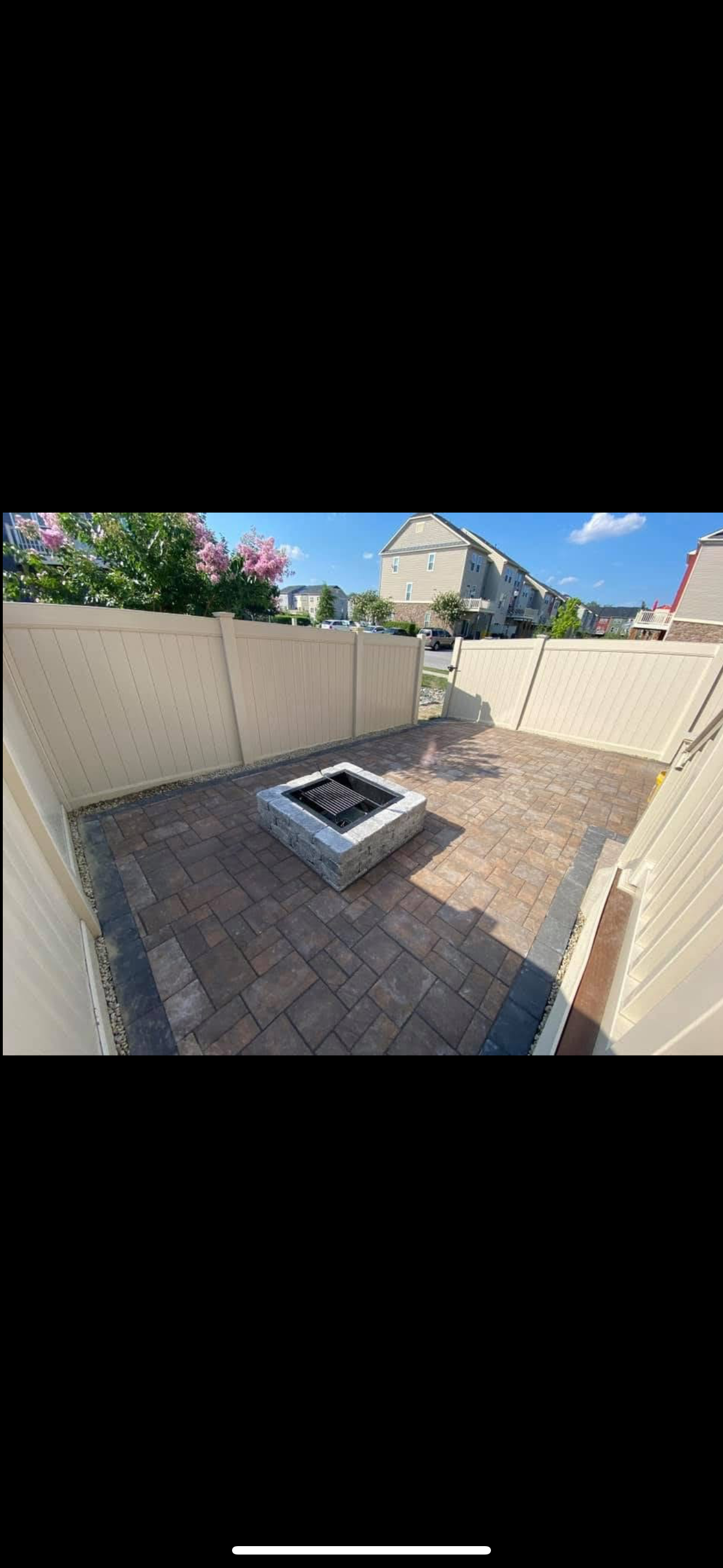 A stone patio featuring a square fire pit in the center, enclosed by tall tan vinyl privacy fencing under a blue sky.