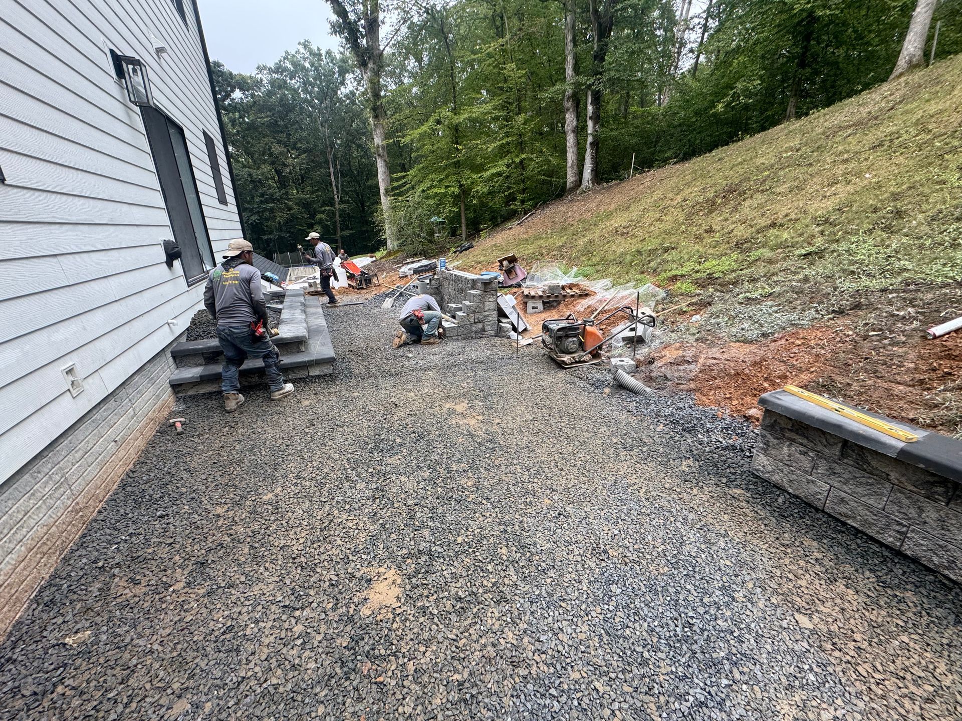 Several construction workers install stone steps and retaining walls on a gravel patio beside a white house.