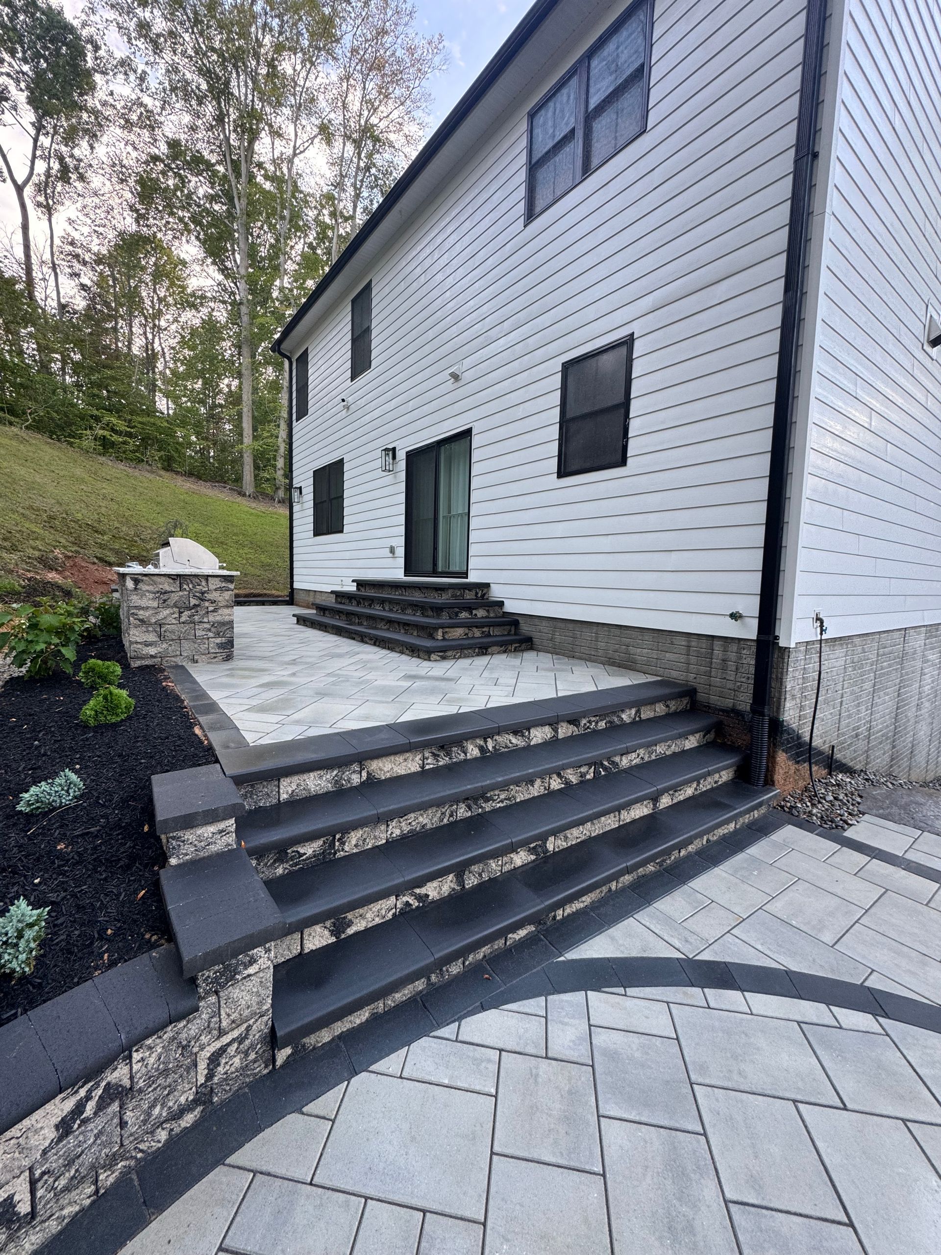 A gray stone patio and stairs lead to a white-sided house with a stone foundation and dark accents.