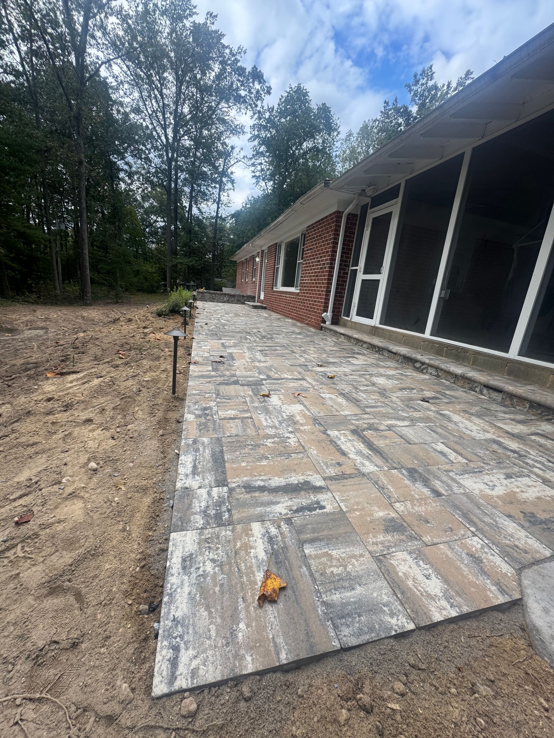 New stone paver patio along the side of a brick house, bordered by bare dirt and trees under a cloudy sky.