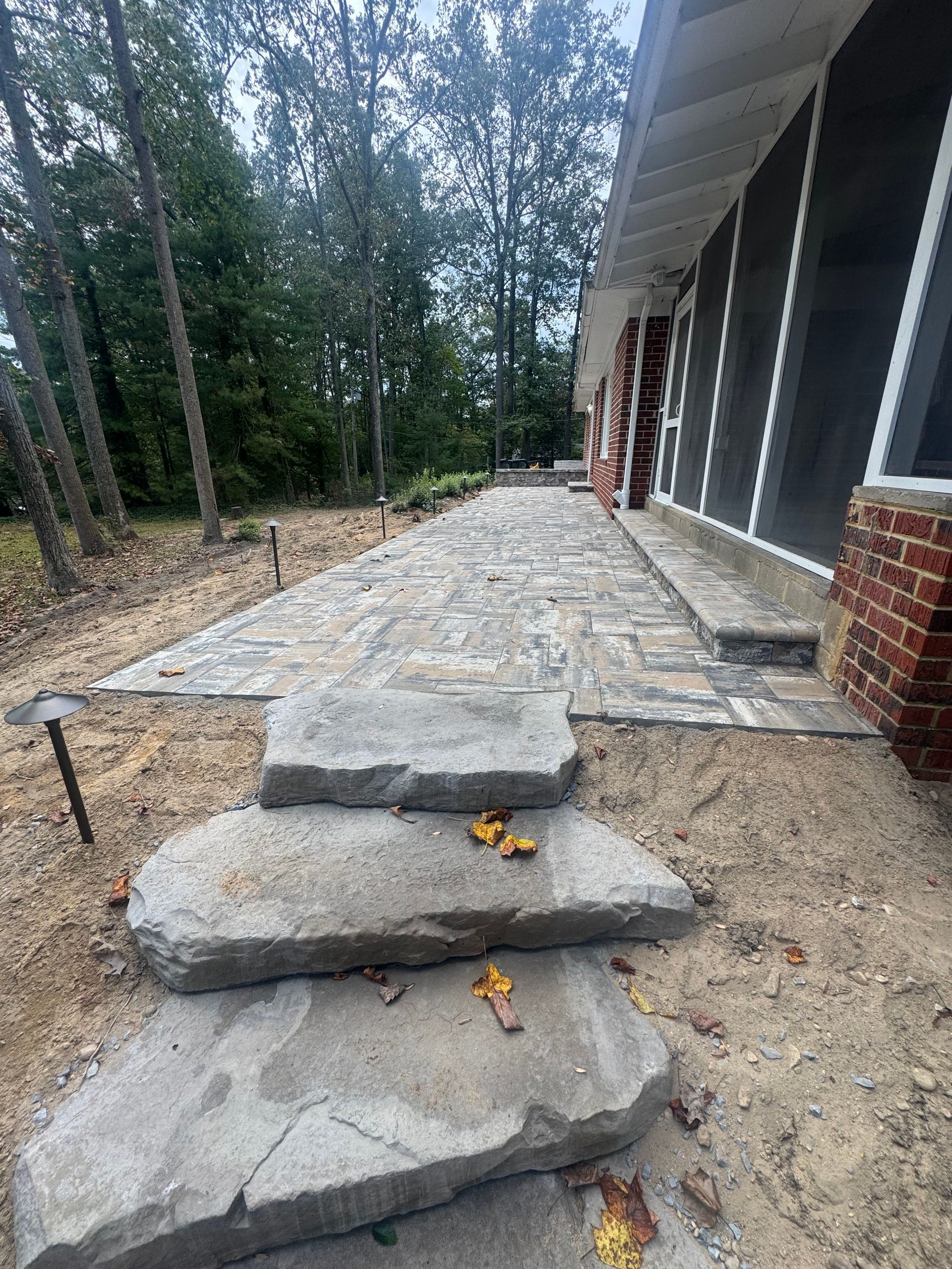 Three natural stone steps lead to a paved patio alongside a brick-walled building exterior surrounded by trees.
