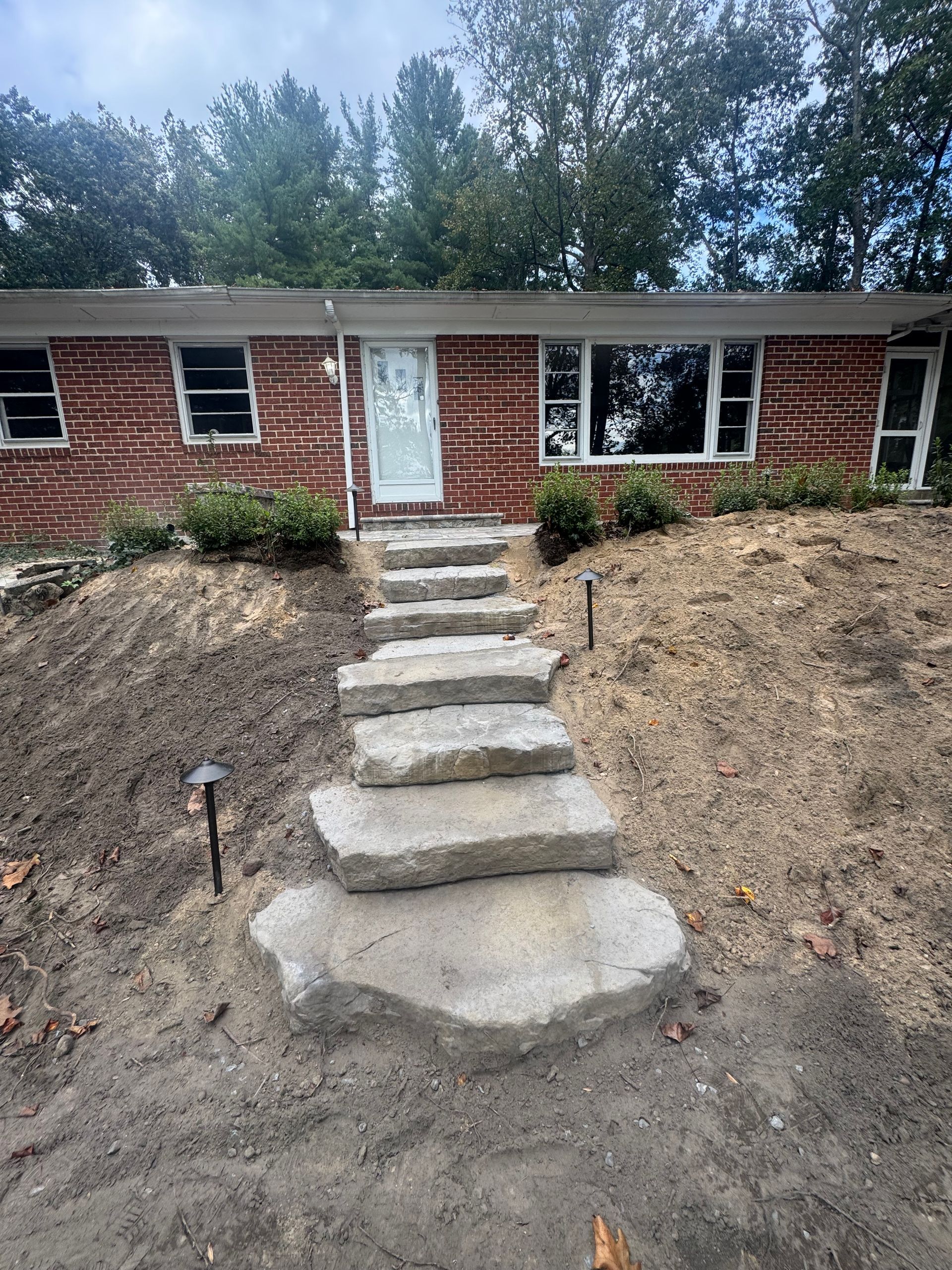 A stone staircase leads up a grassy slope to the front door of a red brick house under a cloudy sky.