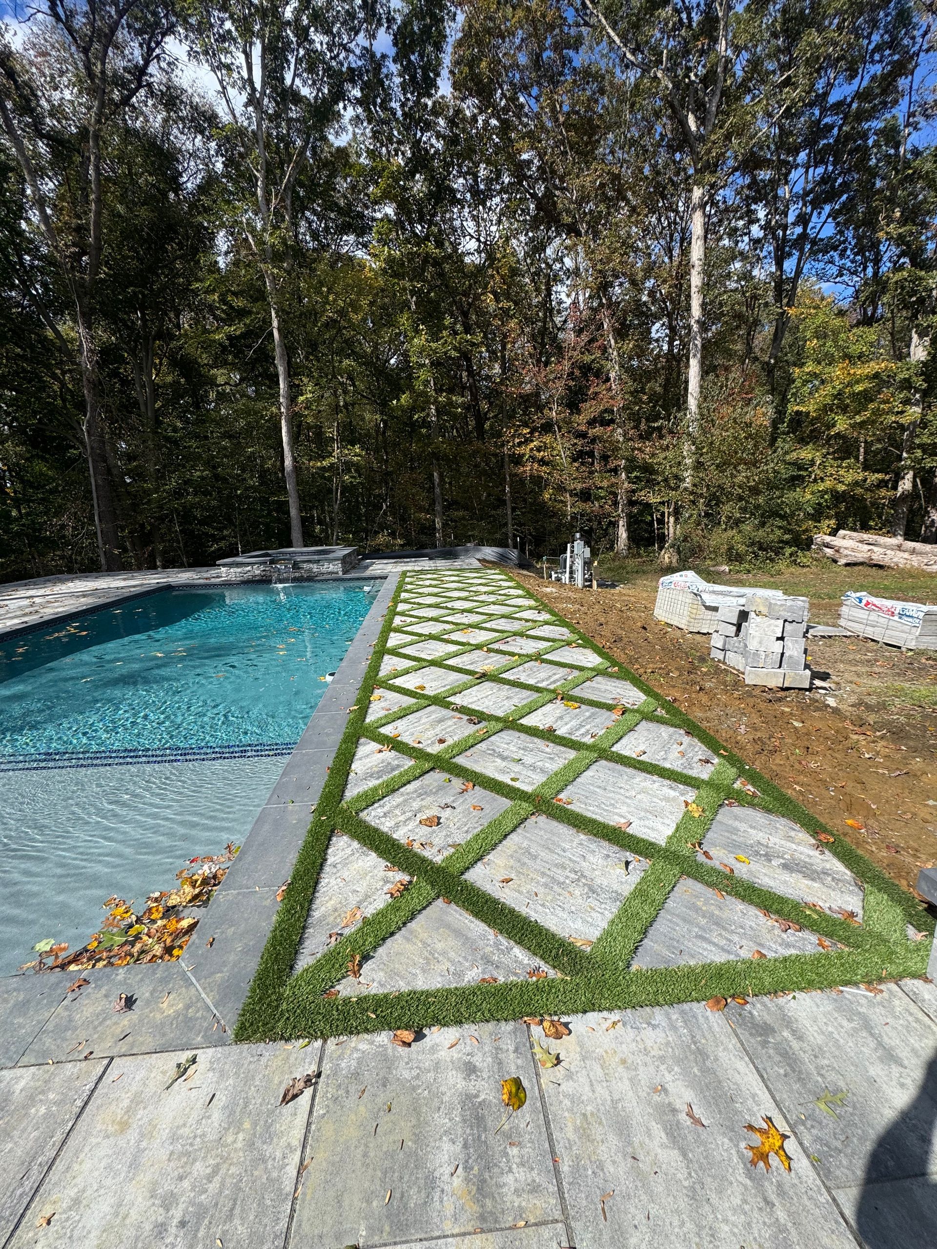 A swimming pool beside a walkway featuring diamond-shaped stone pavers set in green grass, with a forest in the background.