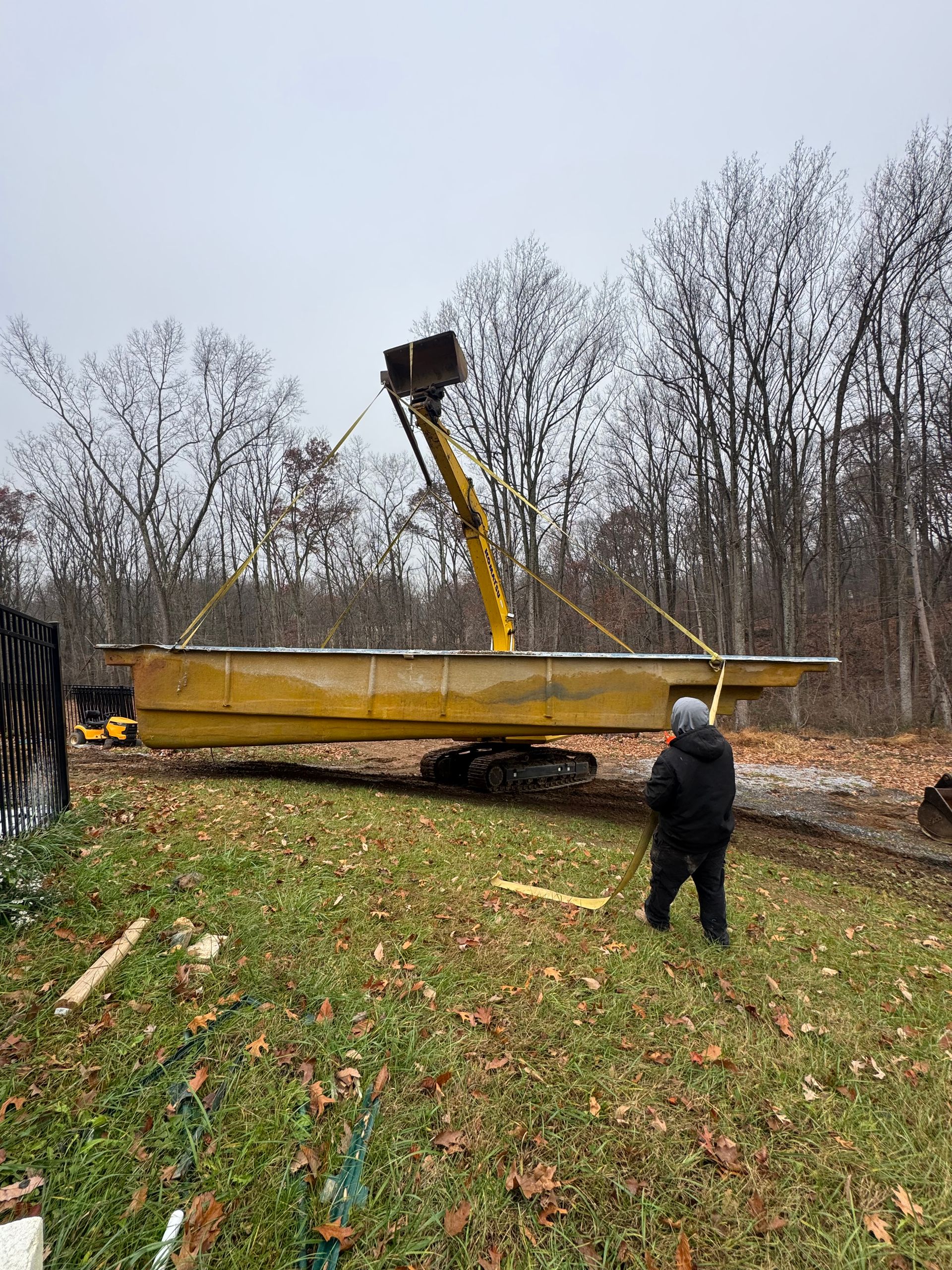 A construction excavator lifts a long, yellow floating dock section in a grassy, wooded yard.