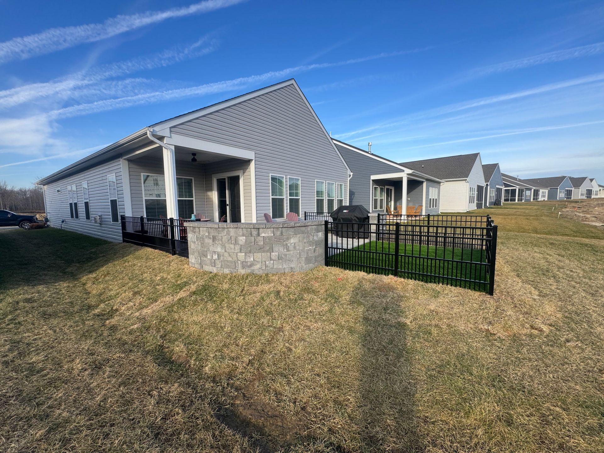 A gray house with a stone patio and black fence, set on a grassy hill under a blue sky with faint airplane trails.