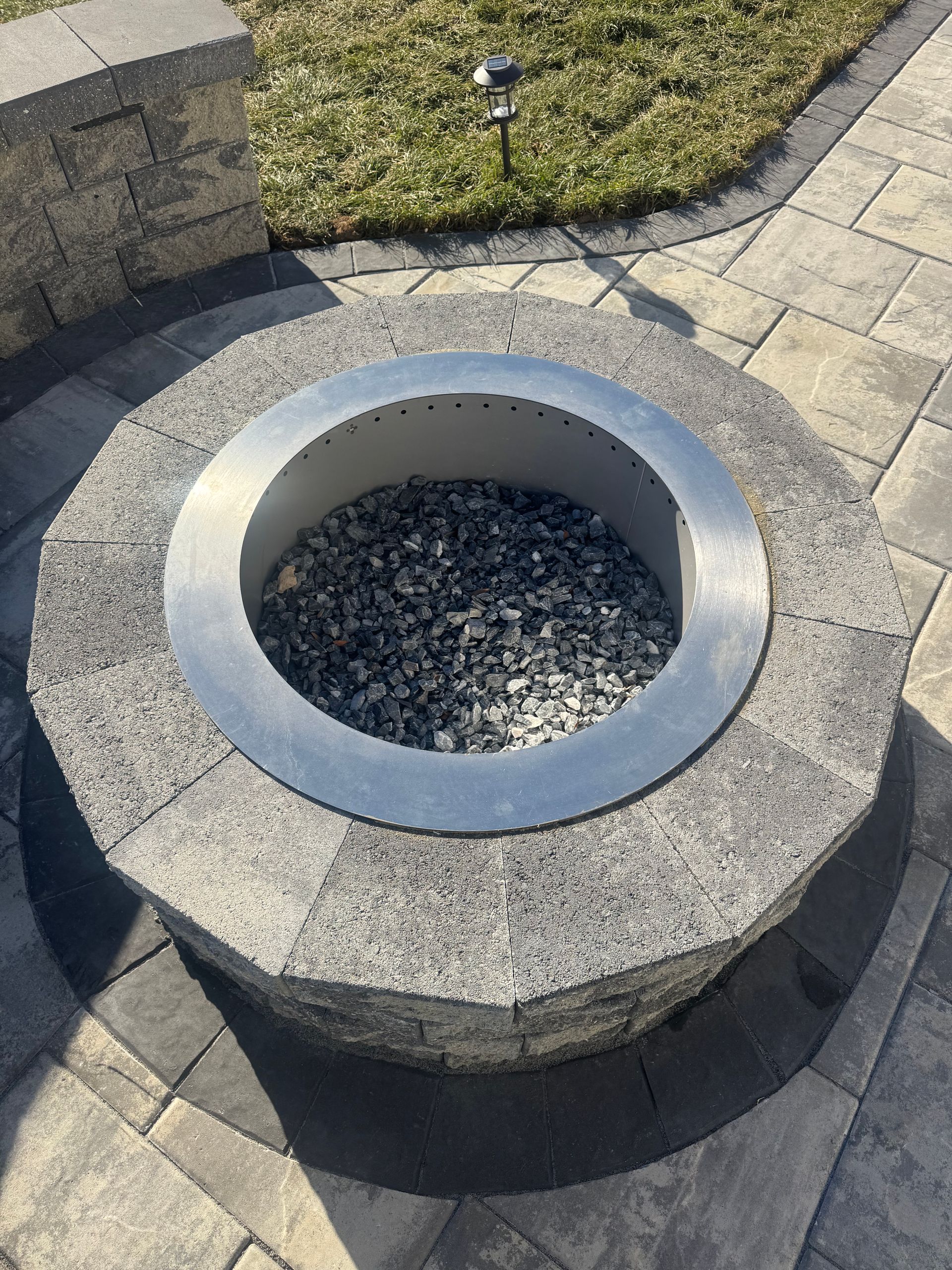 A circular stone fire pit filled with dark gravel, set into a stone patio with a nearby grass lawn.