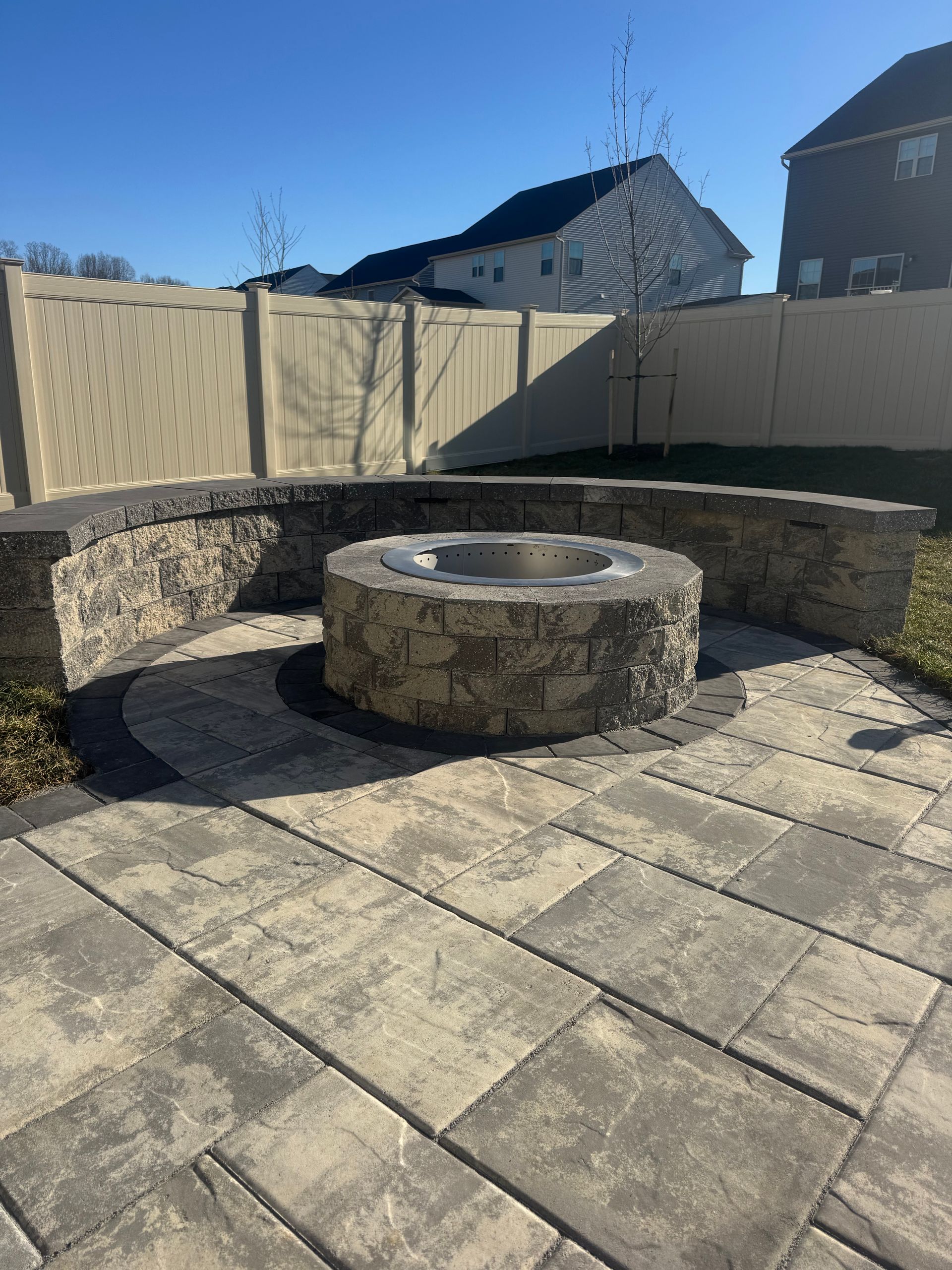 A circular stone fire pit sits on a paved patio in a fenced backyard under a clear blue sky.