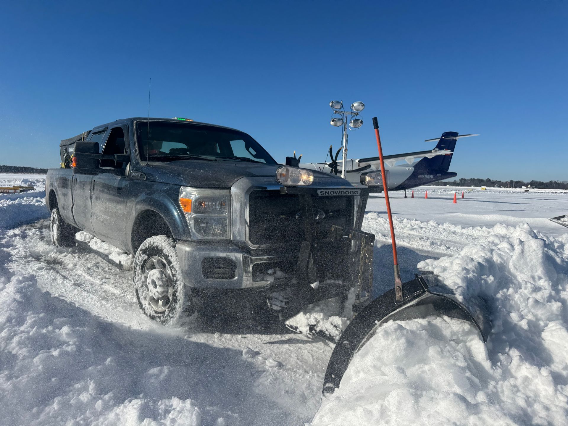 A dark pickup truck with a plow clearing deep snow on an airfield runway near a parked aircraft under a blue sky.