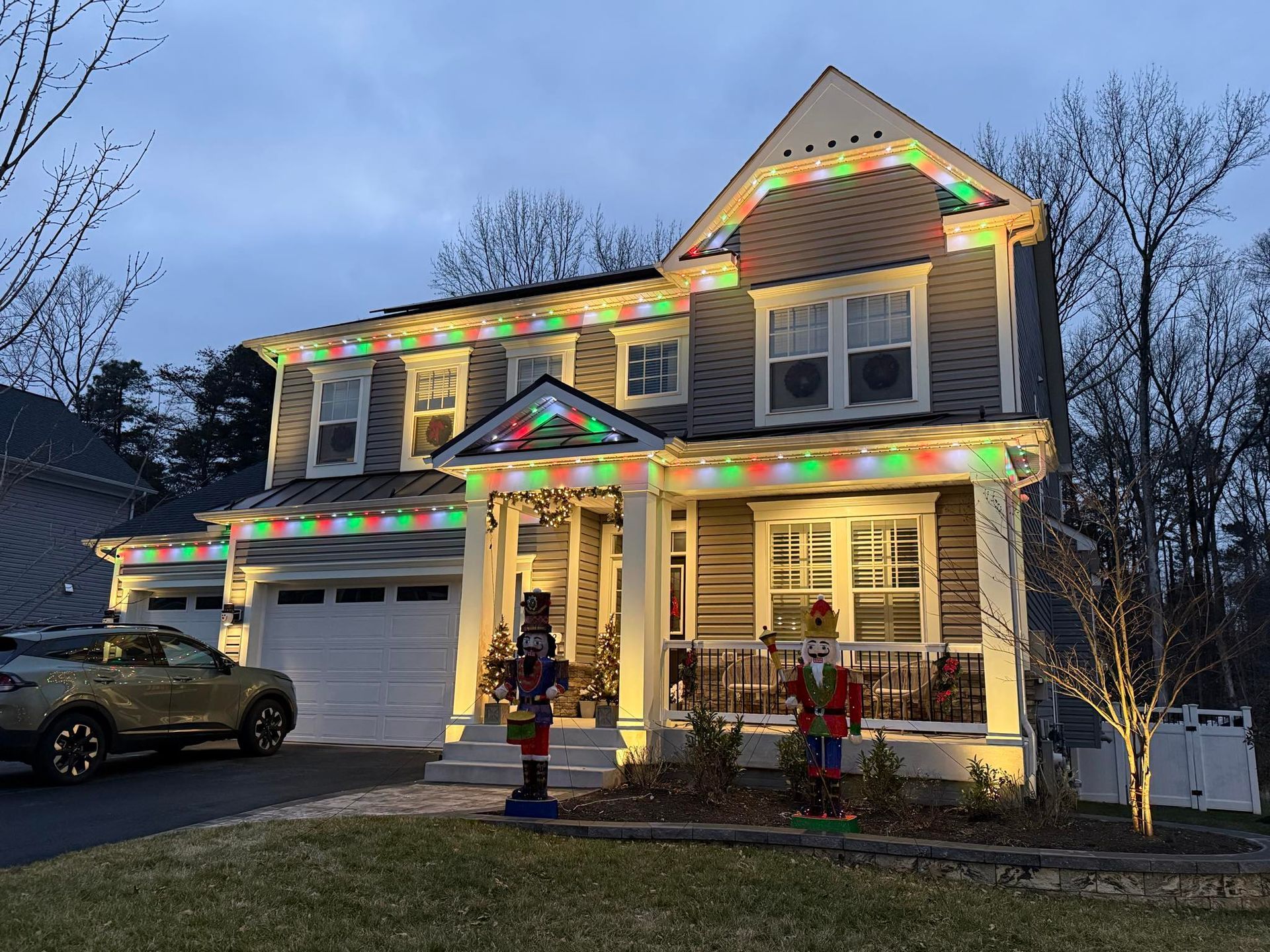 A house exterior at dusk decorated with colorful LED Christmas lights along the rooflines, featuring two large nutcrackers.