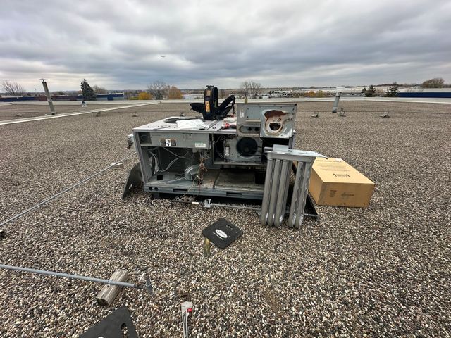 A disassembled HVAC unit sits on a flat, gravel-covered commercial roof next to a cardboard box and some scattered debris.