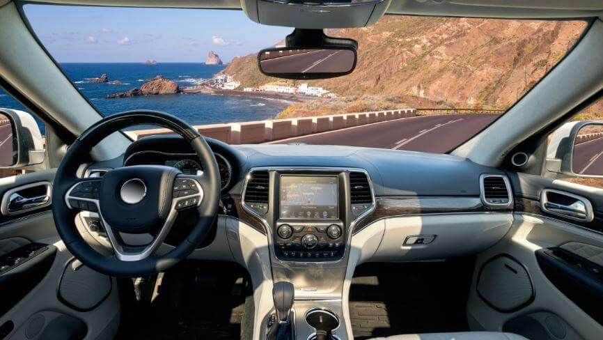 Car interior with steering wheel and dashboard, looking out at a coastal road and ocean through the windshield