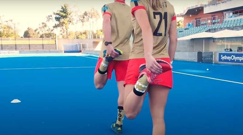 Two people are stretching their legs on a field hockey field.