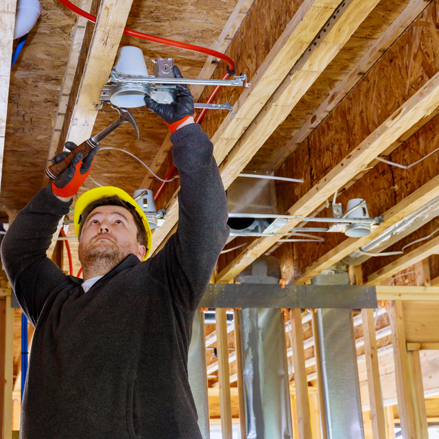 Construction worker installs recessed lighting in a wood-framed ceiling. He wears a hard hat and gloves, working with a hammer.
