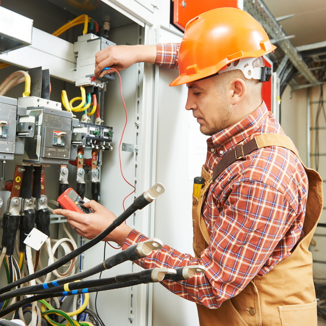 Electrician in orange hard hat, testing wires in an electrical panel.