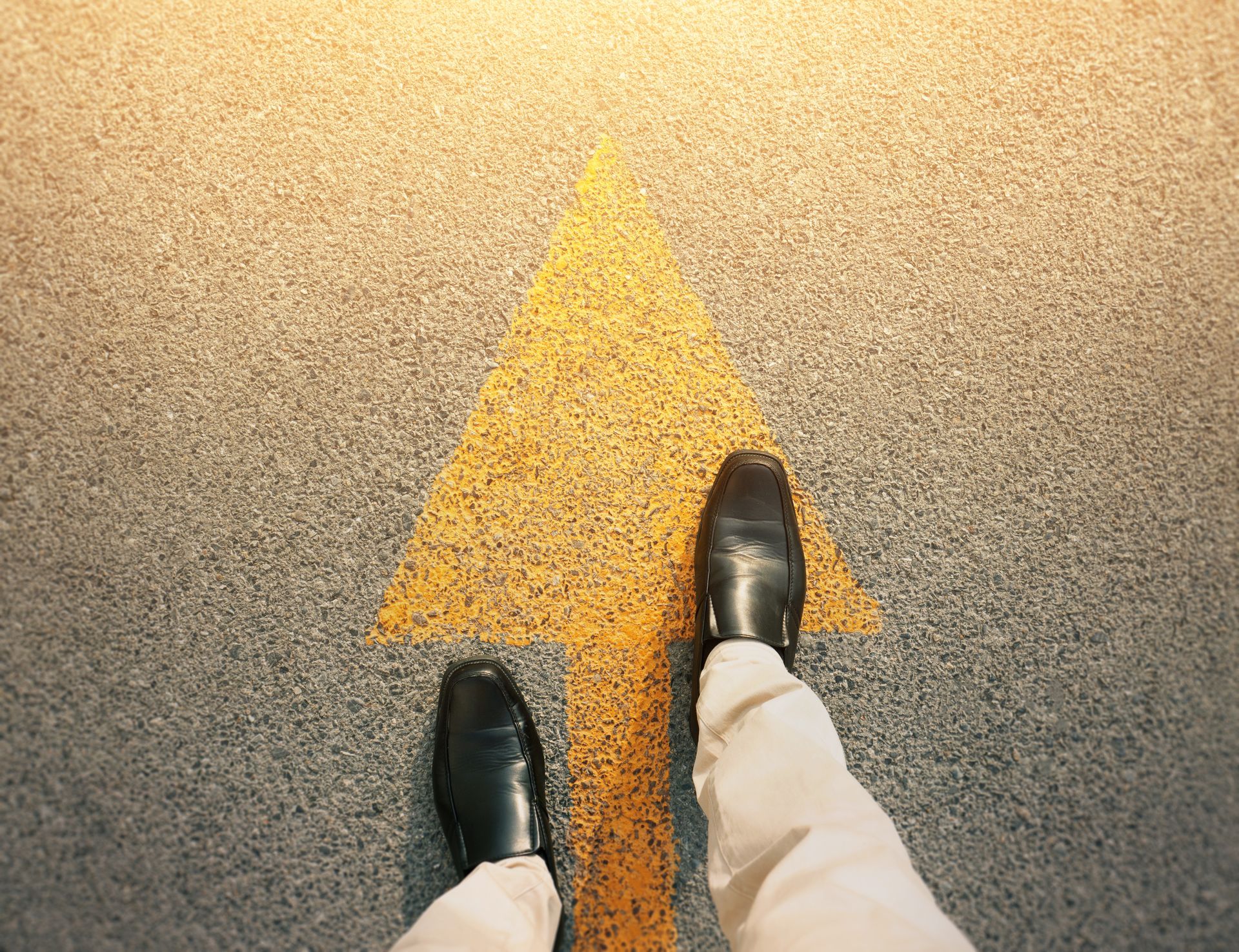 Black shoes walking toward a yellow arrow on pavement, indicating forward movement.