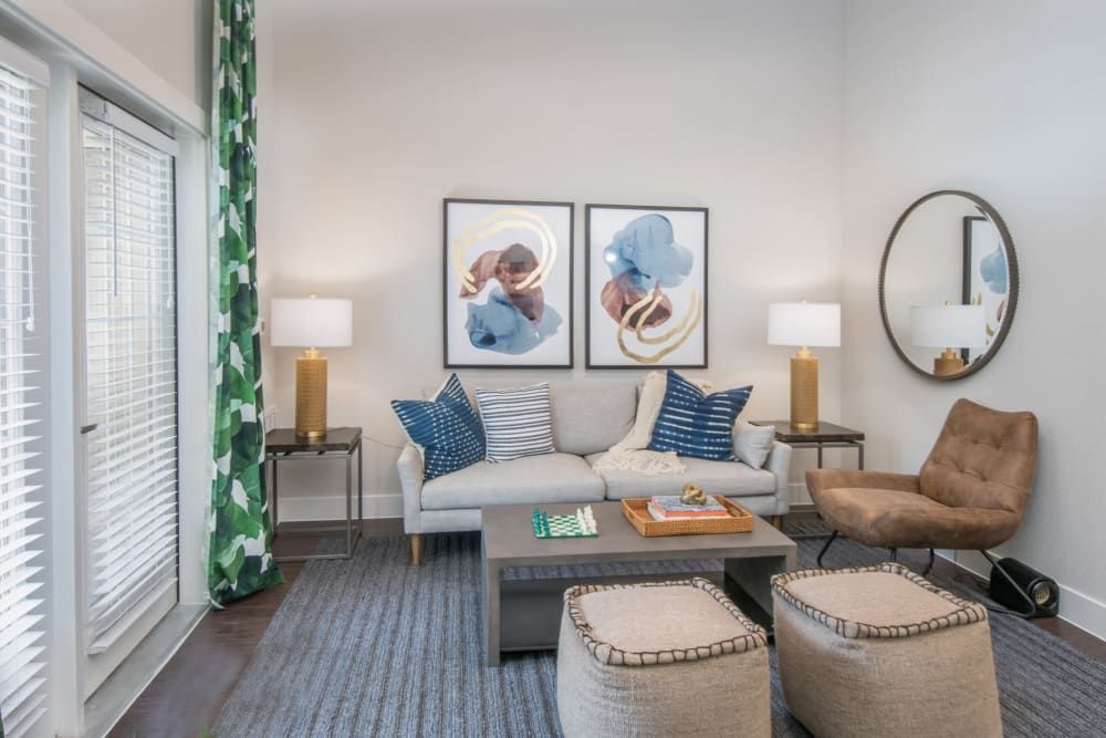 Modern living room interior with a gray sofa adorned with white and blue pillows, flanked by two gold-colored table lamps on side tables. Two abstract art pieces hang above the sofa, opposite a round mirror on the wall. A brown armchair, two beige ottomans, a dark gray rug, and a low wooden coffee table with books and decor items complete the inviting space. Large window with white blinds and green-and-white patterned curtains on the left adds natural light to the room at Marquis Dominion in San Antonio, TX. 