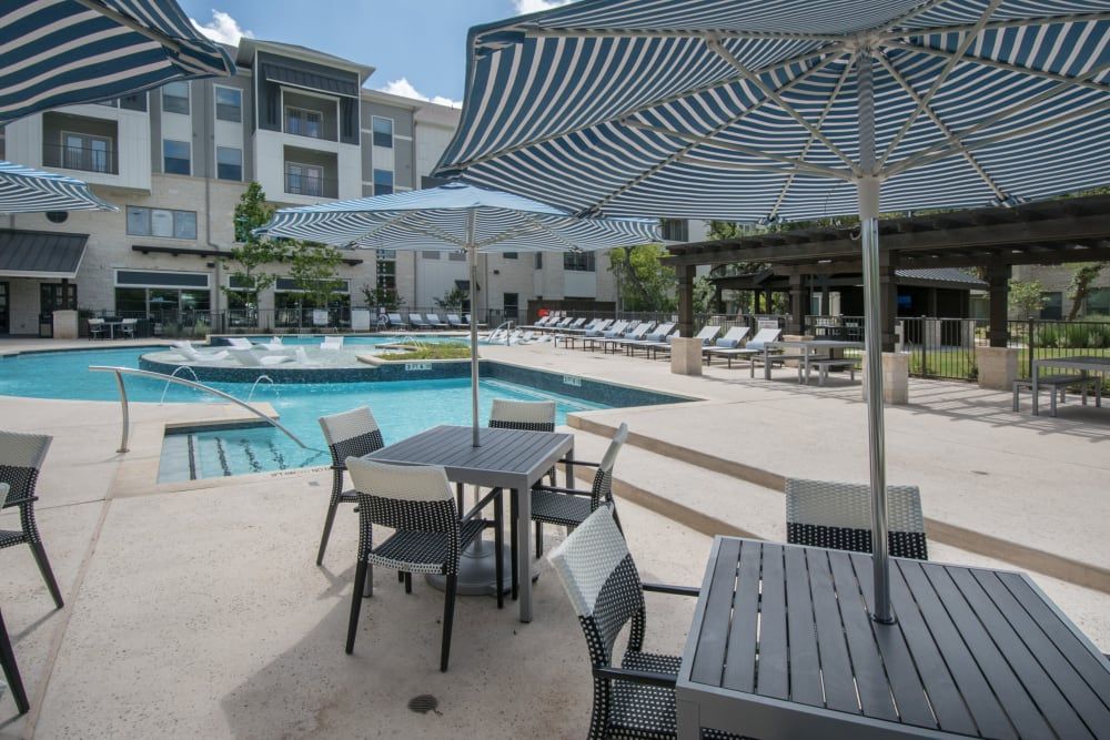 Outdoor swimming pool area at a modern apartment complex with striped umbrellas, patio furniture, and a poolside seating area under a pergola on a sunny day at Marquis Dominion in San Antonio, TX. 