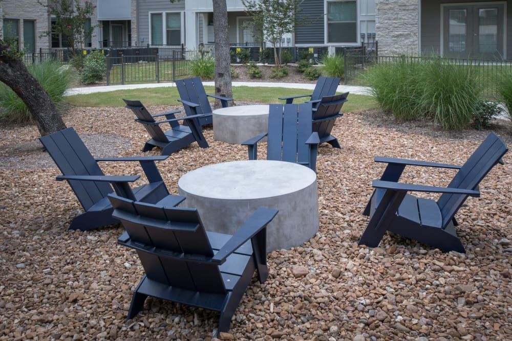 Outdoor relaxation area with modern dark blue Adirondack chairs arranged around a circular concrete table on a bed of small pebbles, with green landscaped grounds and residential buildings in the background at Marquis Dominion in San Antonio, TX. 