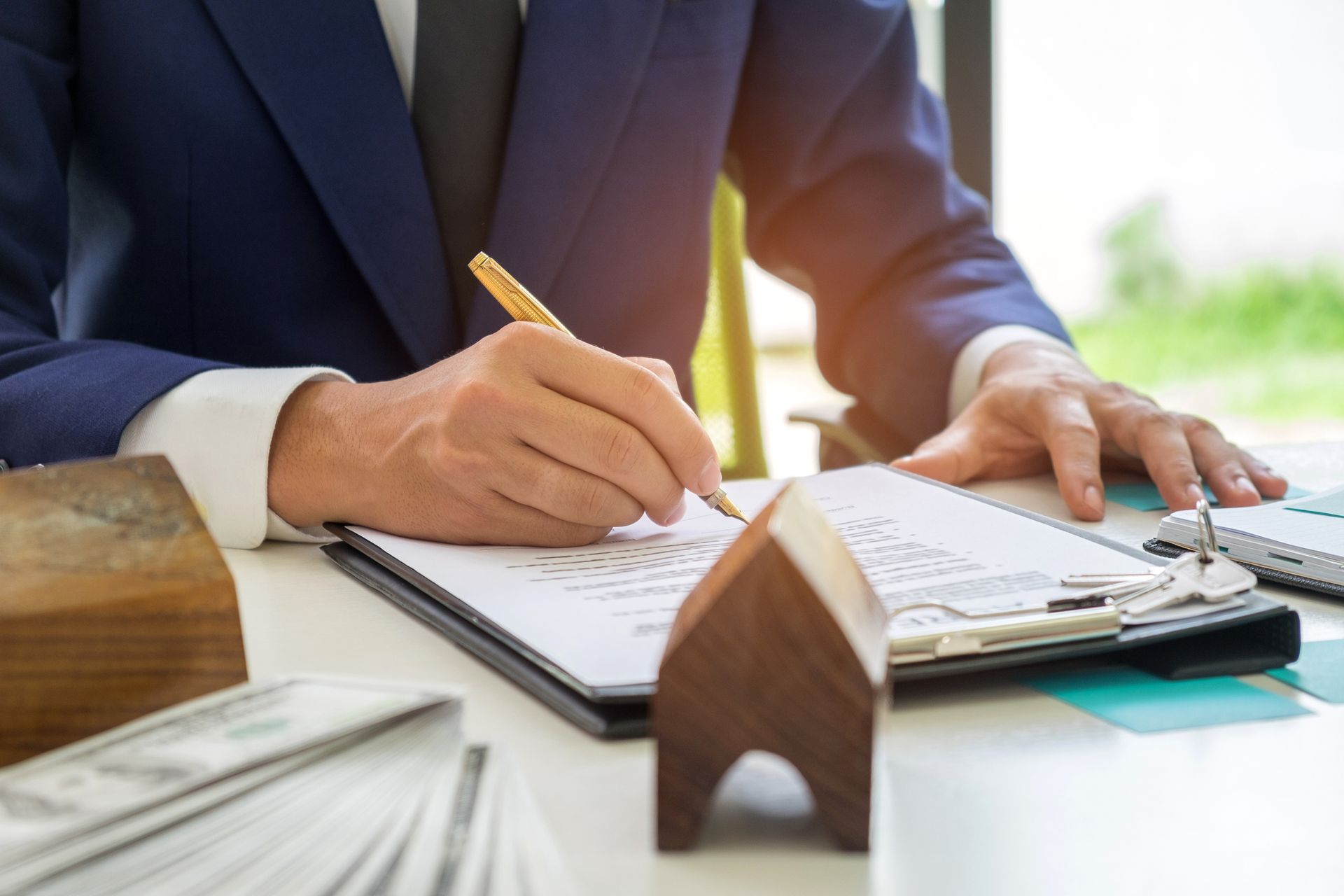 Person in blue suit signing document on a clipboard, miniature house on table.