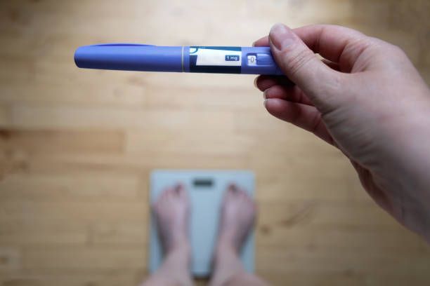 Person standing on a scale bare feet holding a blue GLP-1 injection pen in their hand