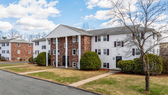 Apartment buildings with brick and white siding, front lawns, and a driveway under cloudy sky
