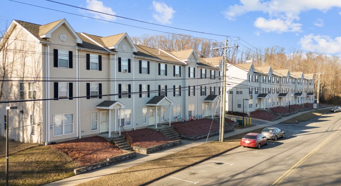 Row of white townhouses on a sunny street with a parked red car and bare trees nearby