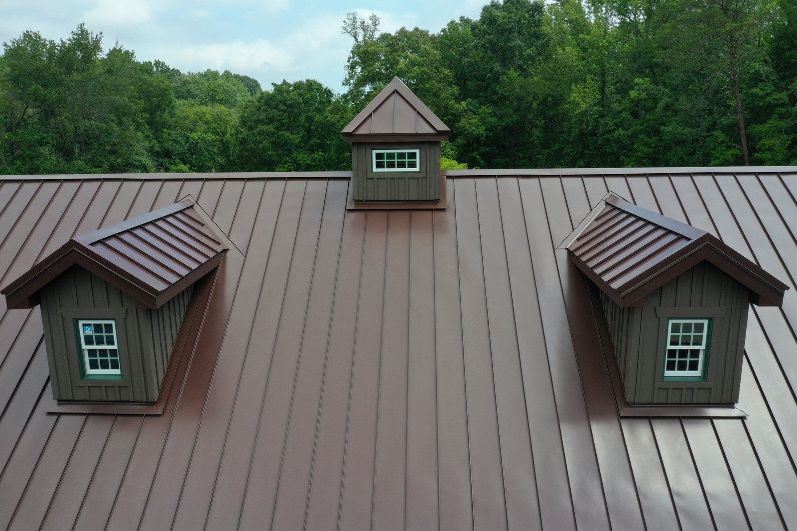 Brown metal roof with three dormers, against a backdrop of green trees.