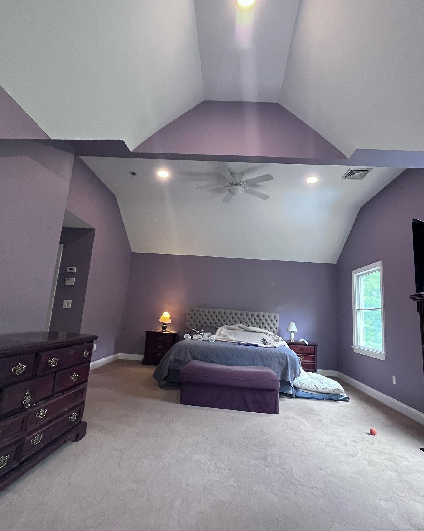 Spacious lavender bedroom with a bed, dresser, lamps, carpet, and a window letting in daylight