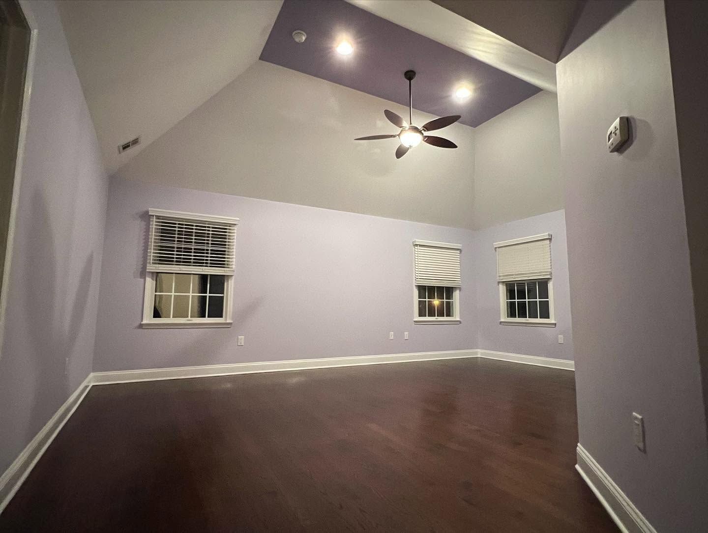 Empty living room with vaulted ceiling, ceiling fan, dark wood floor, and light gray walls with windows.