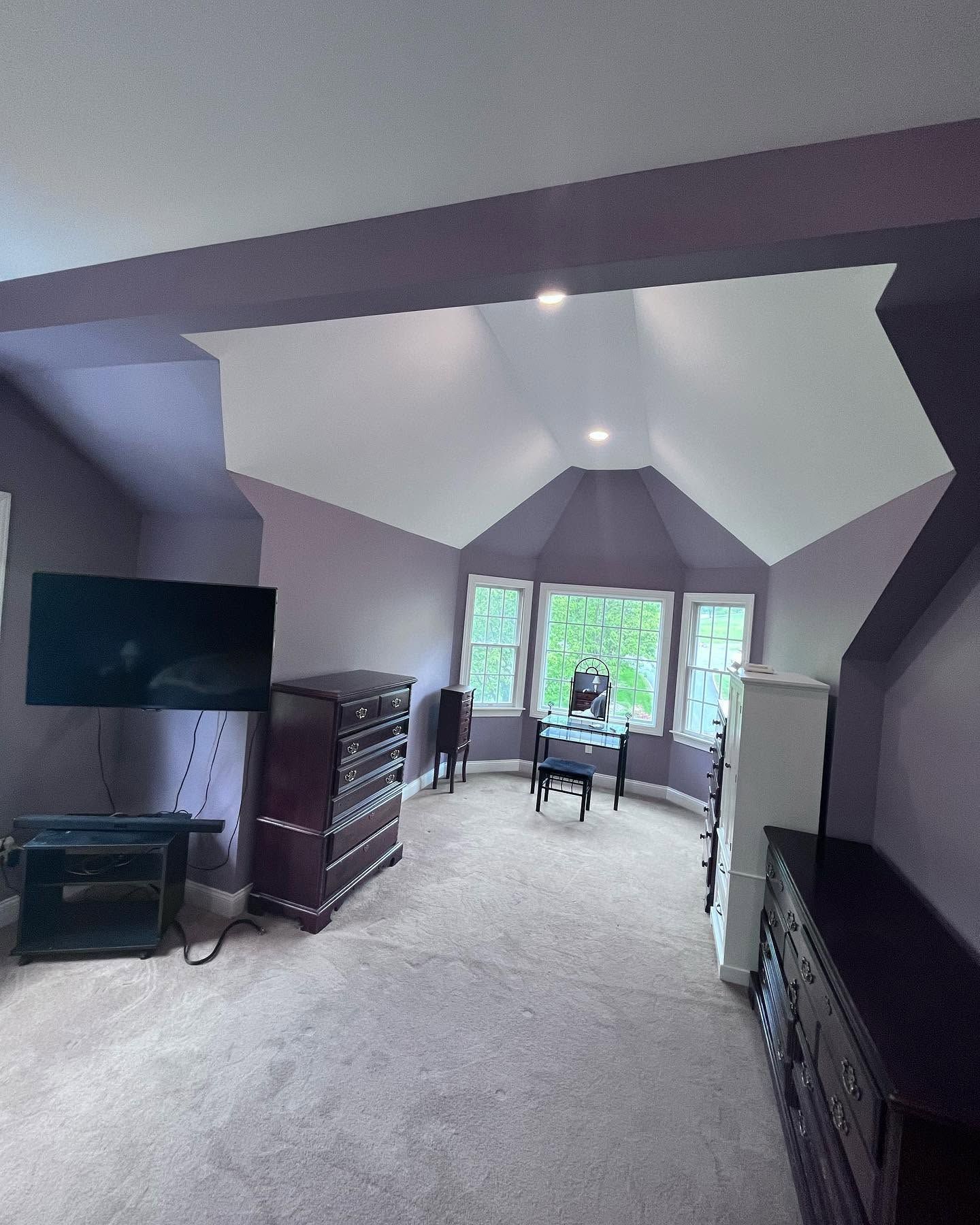 Spacious attic bedroom with vaulted white ceiling, carpet, TV, dresser, and chair by windows.