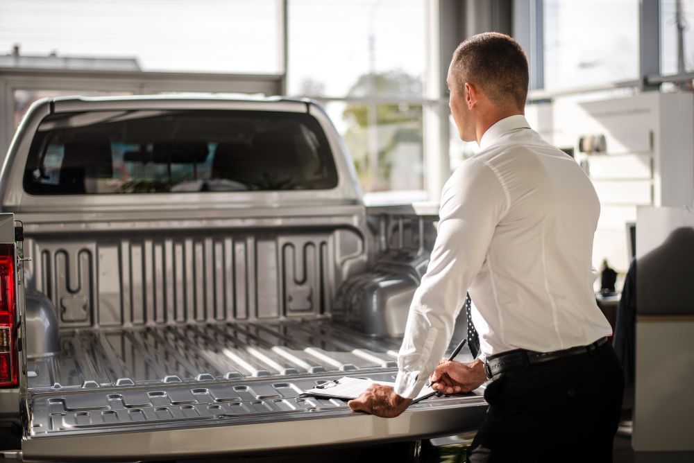 Black SUV Man in white shirt looking into the bed of a silver pickup truck, holding a clipboard in a showroom.